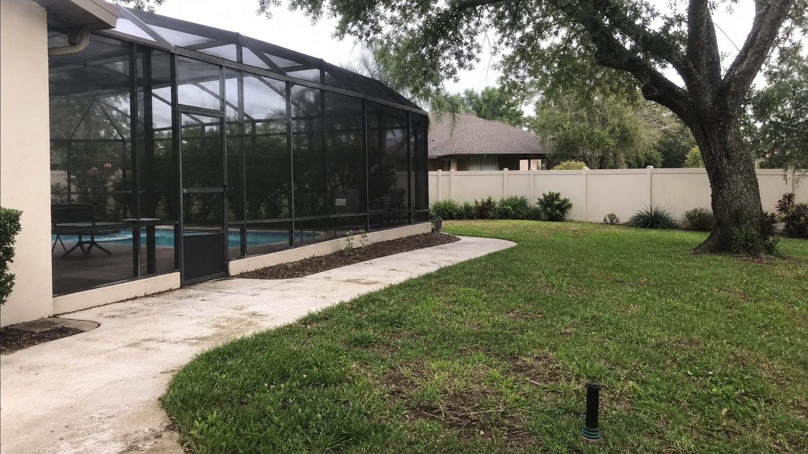 Florida suburban backyard with pool cage, live oak shade, weathered pool deck, irrigation head, overcast afternoon